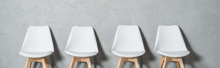 Four white bucket chairs with wooden legs are set against a gray wall, like a waiting room.