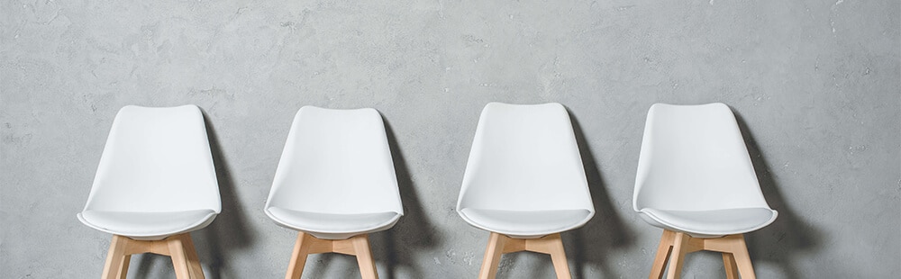Four white bucket chairs with wooden legs are set against a gray wall, like a waiting room.